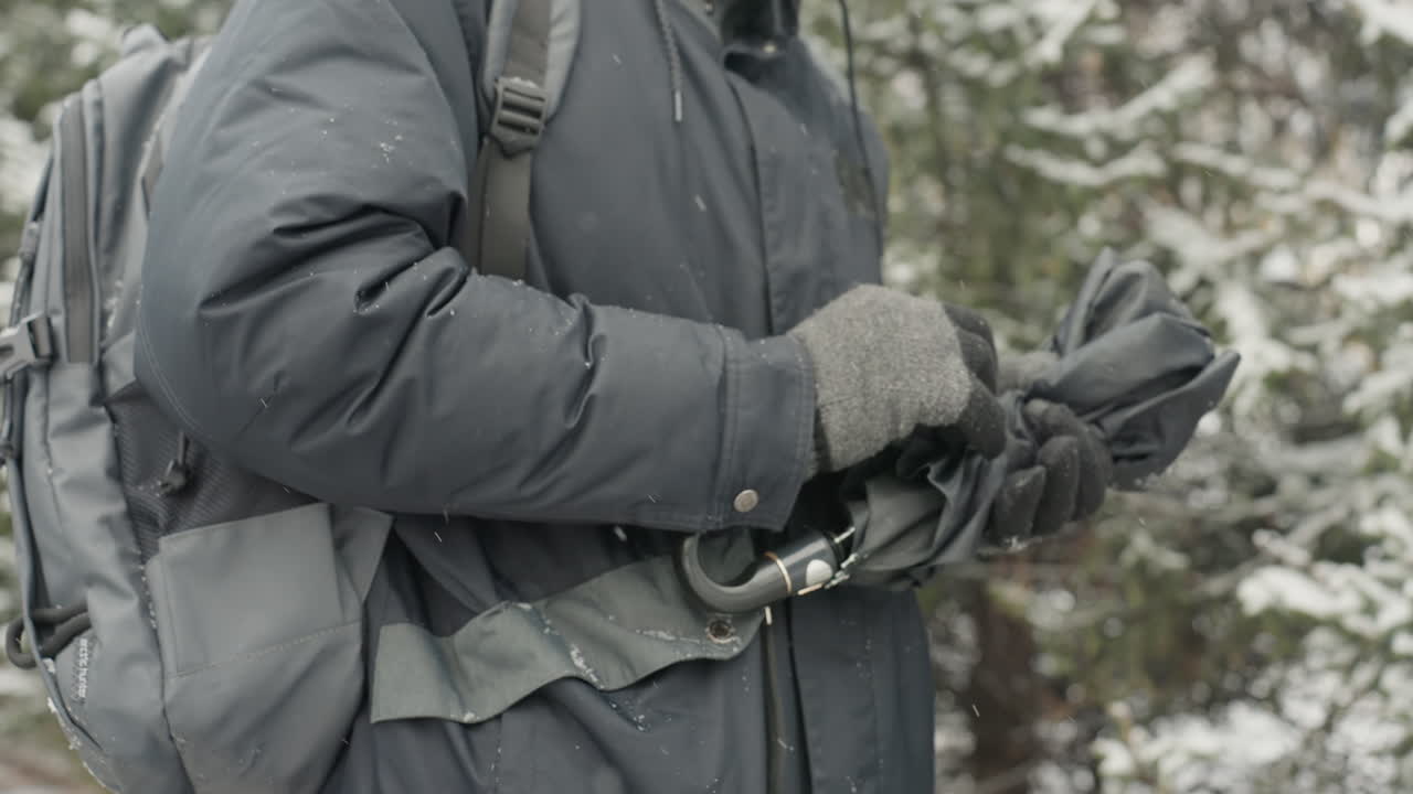 Mann in Winterjacke klappt Regenschirm inmitten schneebedeckter Kiefern zusammen, grauer Rucksack und Strickhandschuhe, Hände fummeln an Verschluss und Speichen, kalter Atem und fallende Flocken, Nahaufnahme von Reißverschluss, Riemen.