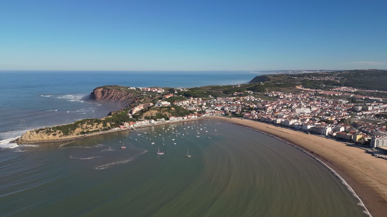 Tomada aérea de drone avançando sobre a baía de São Martinho do Porto, mostrando barcos ancorados, a praia de areia, a cidade costeira e as falésias sob a luz clara da manhã.