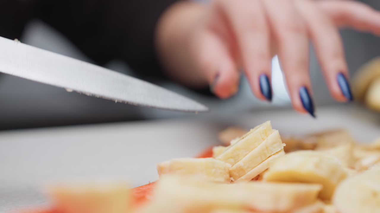 Close up female hands dicing banana with knife on orange rubber board, round slices resting on blade, blurred background, clean counter, fresh fruit prep for snack, manicure with blue nails