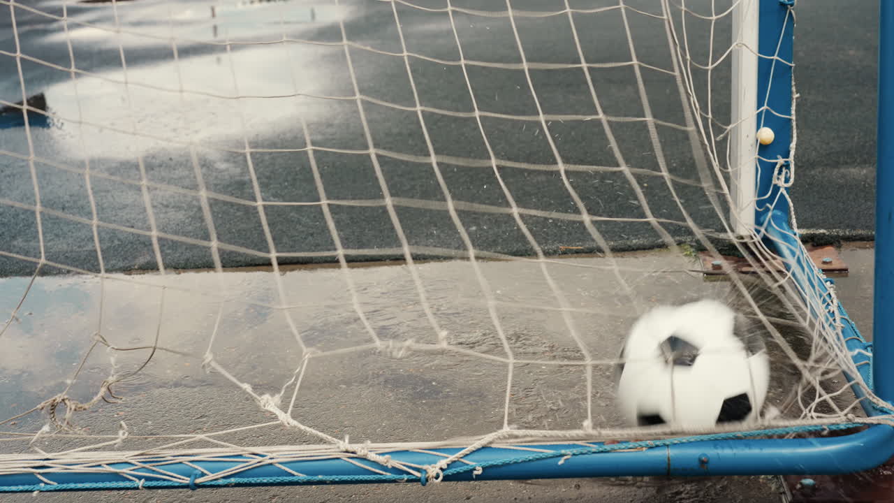 Mavi çerçevede su birikintisi içinde duran futbol topuyla ıslak kale filesi, beton yüzeyde parıldayan yağmur yansımaları, aşınmadan dolayı sarkan file, geç gelen gol sonrası maç sonu dinginliğini çağrıştıran yalnız ve kasvetli atmosfer