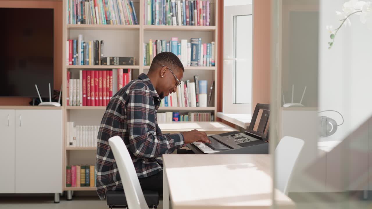 Happy man plays synthesizer at home. Smiling black musician rehearses melody on electrical instrument by bookcase. African American guy performs music