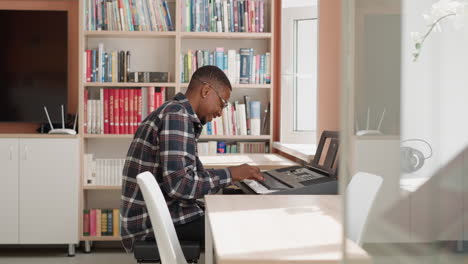 happy man plays synthesizer at home. smiling black musician rehearses melody on electrical instrument by bookcase. african american guy performs music