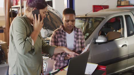 Video-of-two-diverse-female-car-mechanics-using-laptop-and-smartphone,-searching-for-car-parts