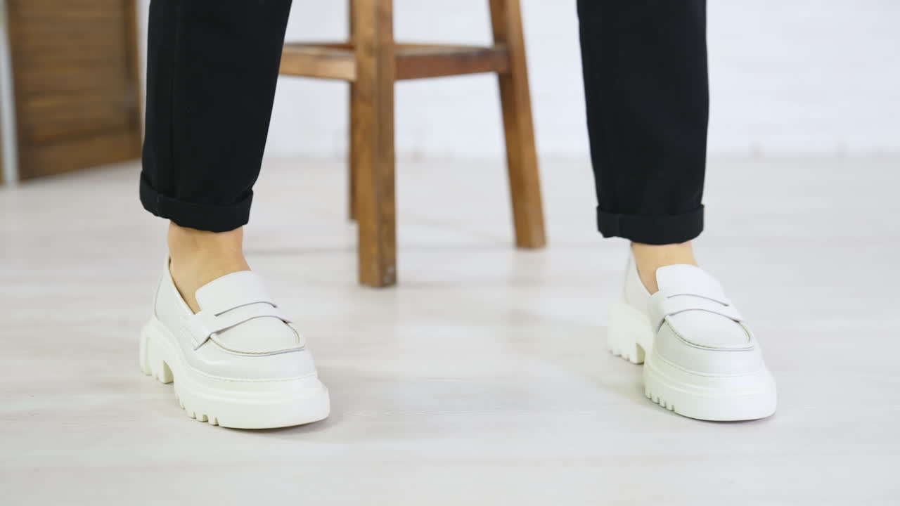 Model wearing black trousers displays white new shoes on massive soles. Close up. Modern footwear presentation at white backdrop.