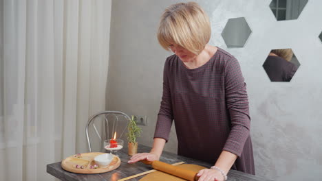 Lady in plaid gown focused on rolling brown dough on kitchen counter, surrounded by candle, wooden tray, and chair, with a wall mirror reflecting her action