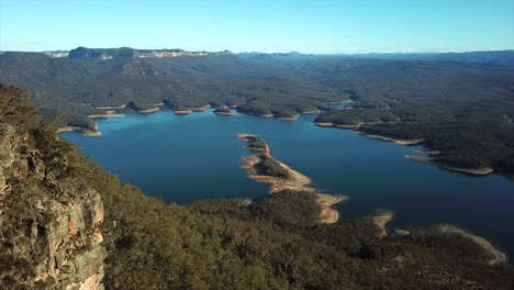 aerial: drone flying next to a mountain range towards a beautiful blue lake in new south wales, australia