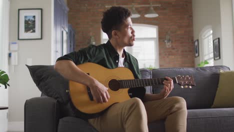 happy biracial man sitting on sofa in living room playing guitar, singing