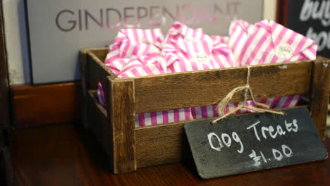 a rustic wooden crate filled with pink-striped paper bags labeled "dog treats" on a counter, with a chalkboard sign pricing