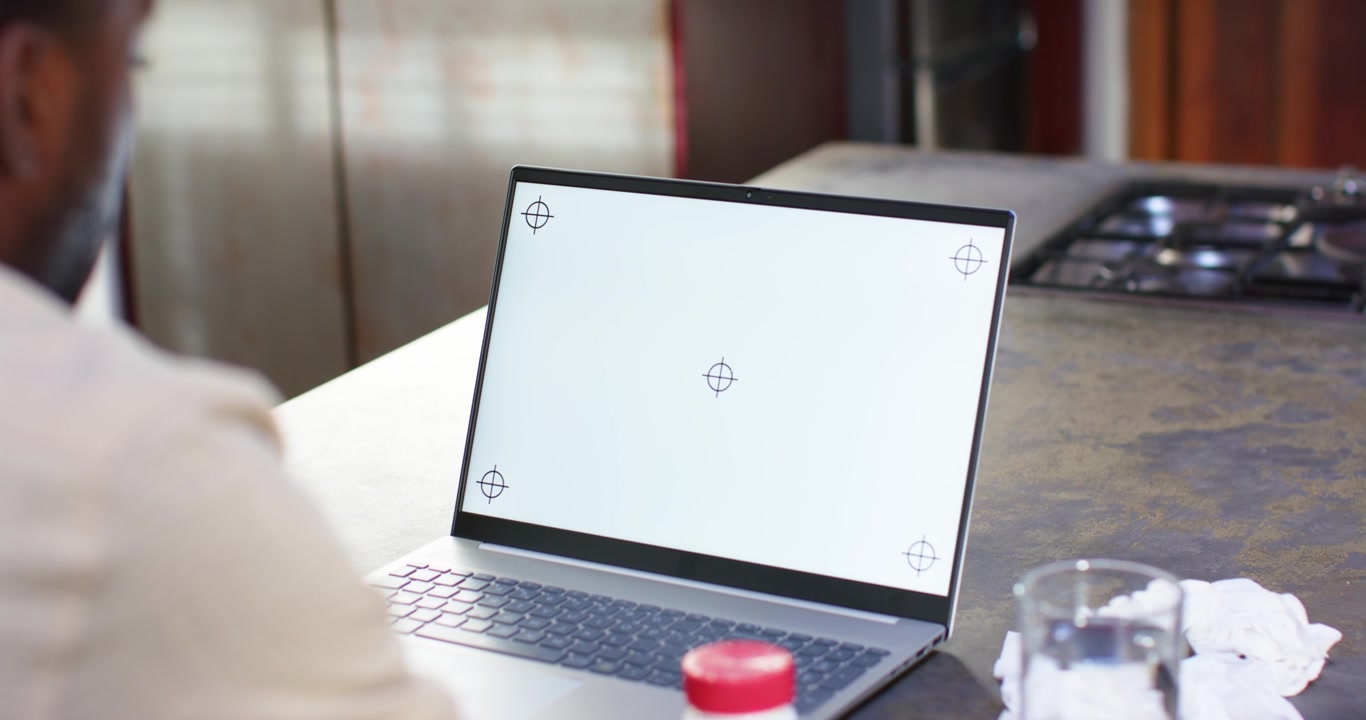 Man sitting at kitchen counter watching blank laptop screen with targets, adjusting for webcam