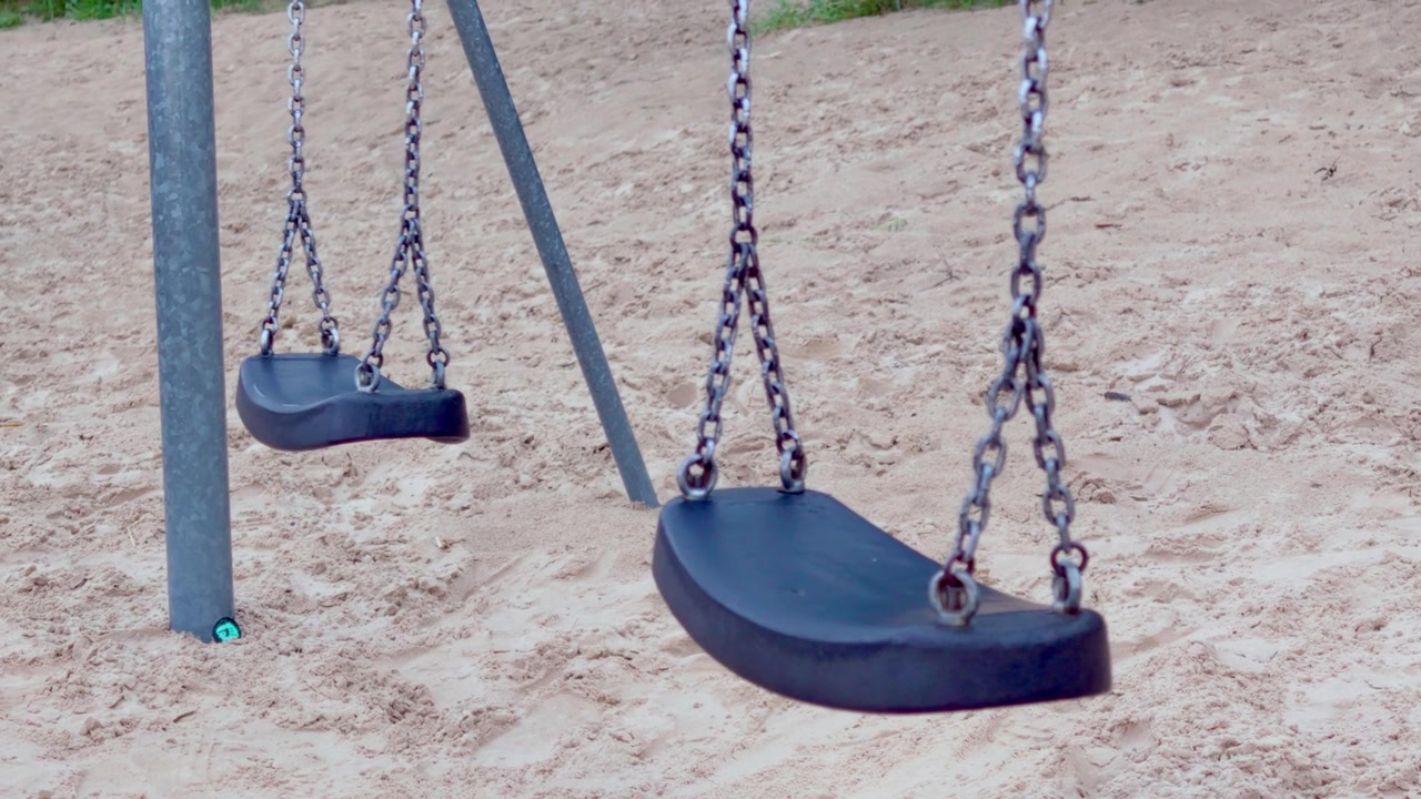 Inquadratura media di altalene nere vuote di un parco giochi che oscillano dolcemente nel vento sopra sabbia pulita vicino alla spiaggia a Jurmala, evocando un'atmosfera nostalgica di svago infantile