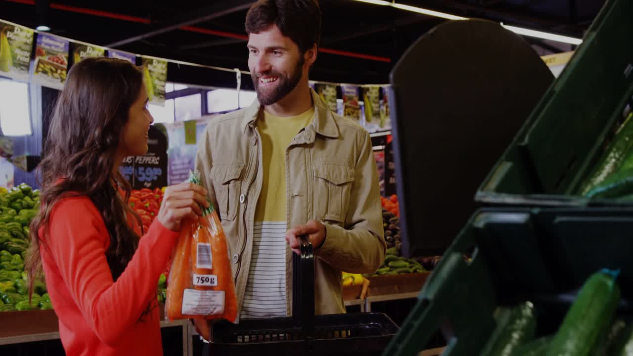 Woman picking up bag of carrots, checking label while grocery shopping, man holding basket steady
