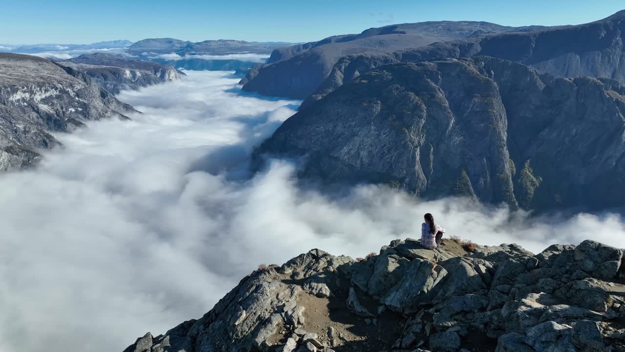 Drone slowly orbits a woman sitting on Bakkanosi cliff with panoramic view of the fjord and mountains. She turns head, showing a ponytail in slow motion