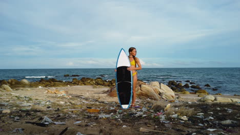 young sexy female surfer in bikini holding surfboard on polluted ocean beach filled with plastic waste and trash, global warming climate change concept