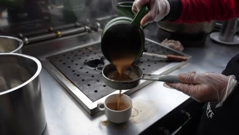 gloved hand pours spiced tea through a strainer into a cup at a street-side setup