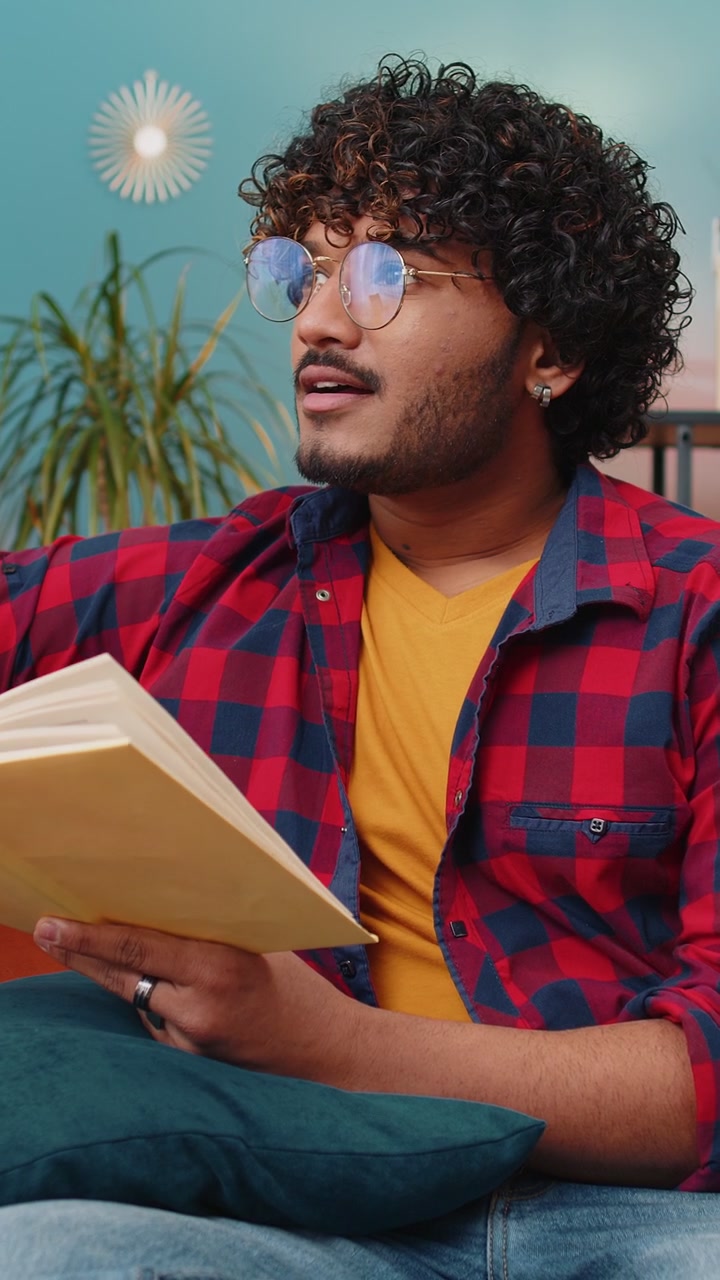 Indian man reading interesting book turning pages smiling enjoying literature taking a rest on sofa