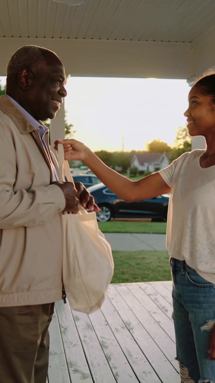 Vertical video: Extending teen in white tee, jeans handing tote for grandpa to carry on porch