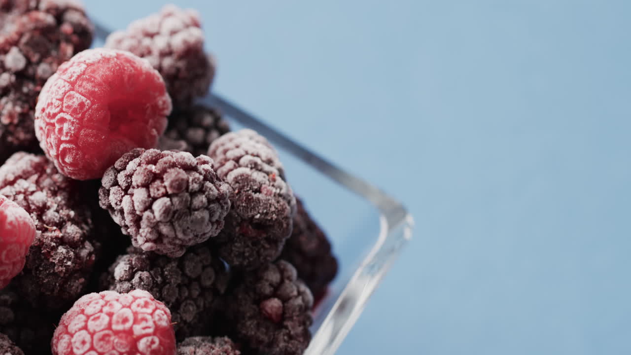 Micro video of close up of raspberries in glass bowl with copy space on blue background