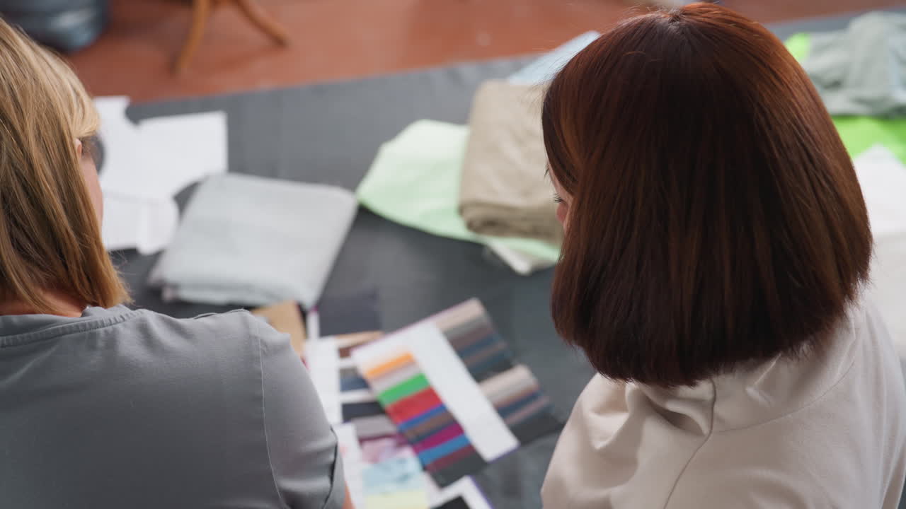 Two seamstress studying fabric color pallet on table, discussing textile options, creative workspace filled with cloth samples and team brainstorming design ideas for fashion production