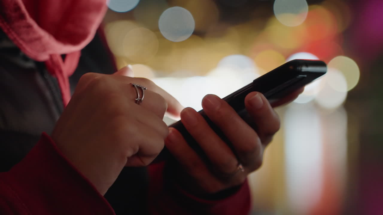 Primo piano di mani che tengono uno smartphone di notte, mani con felpa rossa e sciarpa toccano lo schermo contro le luci bokeh sfocate della città, anelli sulle dita creano un'intima scena serale di messaggi con un morbido bagliore al neon