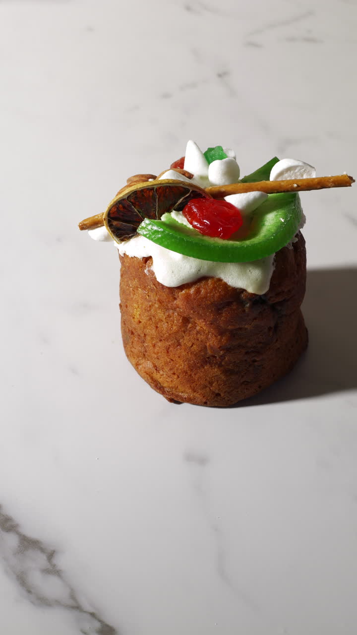 Close-up of a hand placing a festive cupcake topped with whipped cream, dried fruit, jelly candy, and a pretzel stick on a marble surface. The scene is brightly lit and showcases detailed textures.