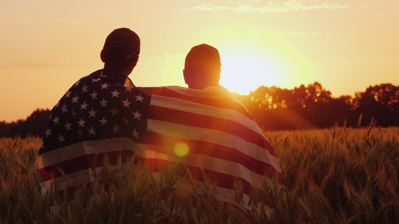 un hombre y su hijo admiran la puesta de sol sobre un campo de trigo envuelto en la bandera de los estados unidos