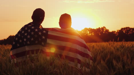 Ein-Mann-Und-Sein-Sohn-Bewundern-Den-Sonnenuntergang-über-Einem-Weizenfeld,-Das-In-Die-Flagge-Der-Usa-Gehüllt-Ist