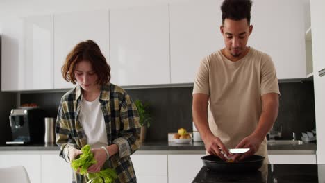 Un-Joven-Feliz-De-Piel-Negra-Con-Una-Camiseta-Beige-Prepara-Huevos-Revueltos-Para-El-Desayuno-Junto-Con-Su-Joven-Novia-Adulta-De-Cabello-Castaño-Y-Una-Camisa-A-Cuadros-Verde-Que-Prepara-Una-Ensalada-En-Una-Cocina-Moderna-En-Un-Apartamento-Por-La-Mañana.