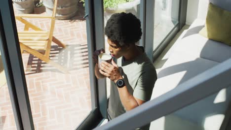 hombre afroamericano mirando por la ventana y bebiendo café en casa