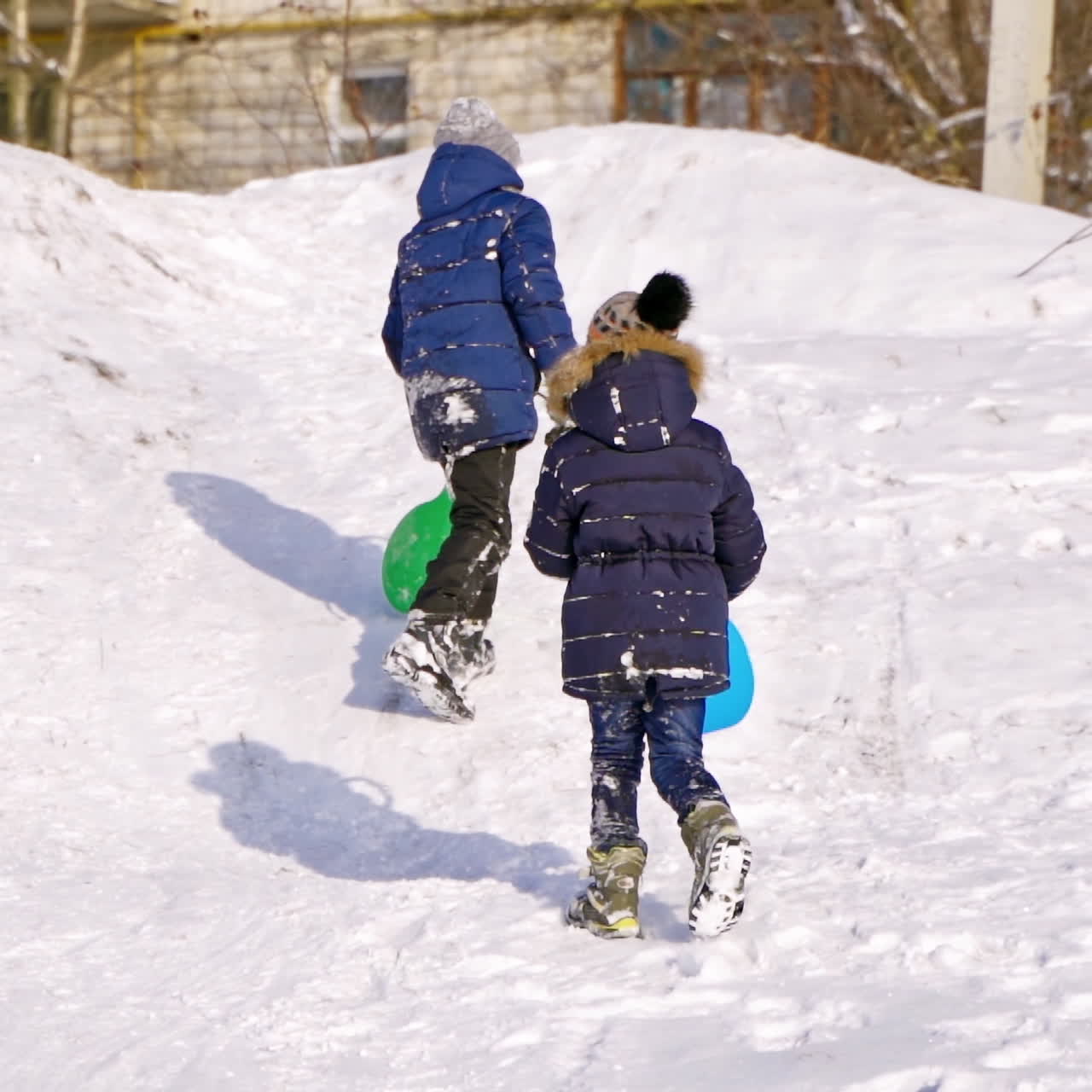 Children having fun, playing and laughing on snowy winter walk in nature. Frost winter season. Cute kids with plastic sledges walk up a hill outdoors on winter day. Winter games and fun