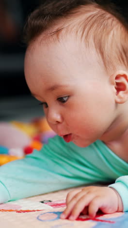 cute child with funny hairdo reaches the edge of a mat he is lying at. lovely baby is interested with pictures at the cover. vertical video
