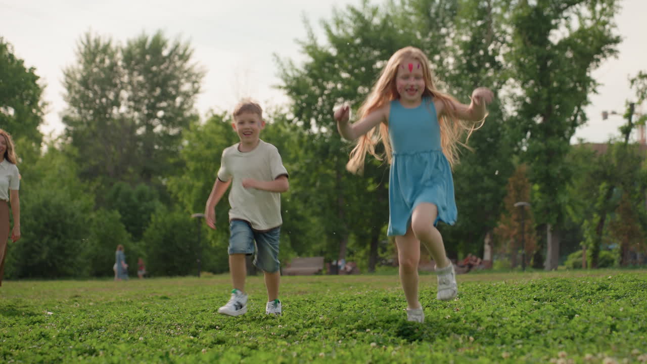 Two children racing playfully on grass in park while woman watches in ...