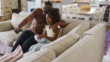African-american-daughter-and-her-father-on-couch-eating-popcorn-and-laughing-at-tv
