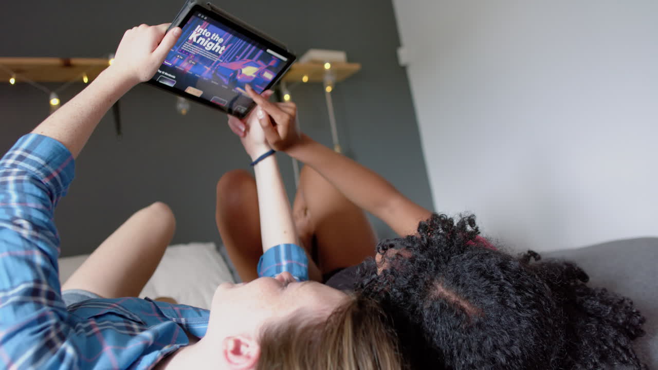 amigas adolescentes felices y diversas acostadas en la cama y usando tableta con película en casa, en cámara lenta