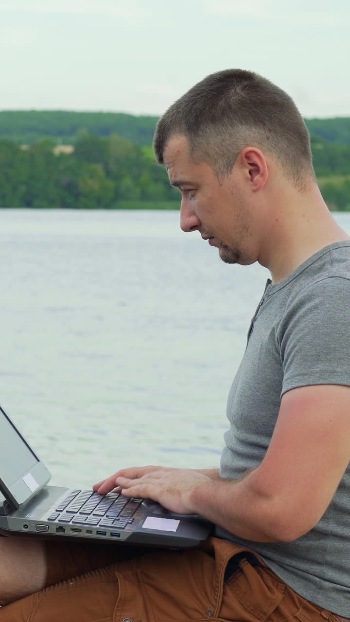 Handsome man sitting on a wooden pier with laptop. Vertical video