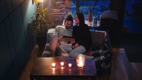 romantic evening: a brunette guy and a girl are sitting on the couch and looking at the screen of the tablet in the evening. there are candles on the table