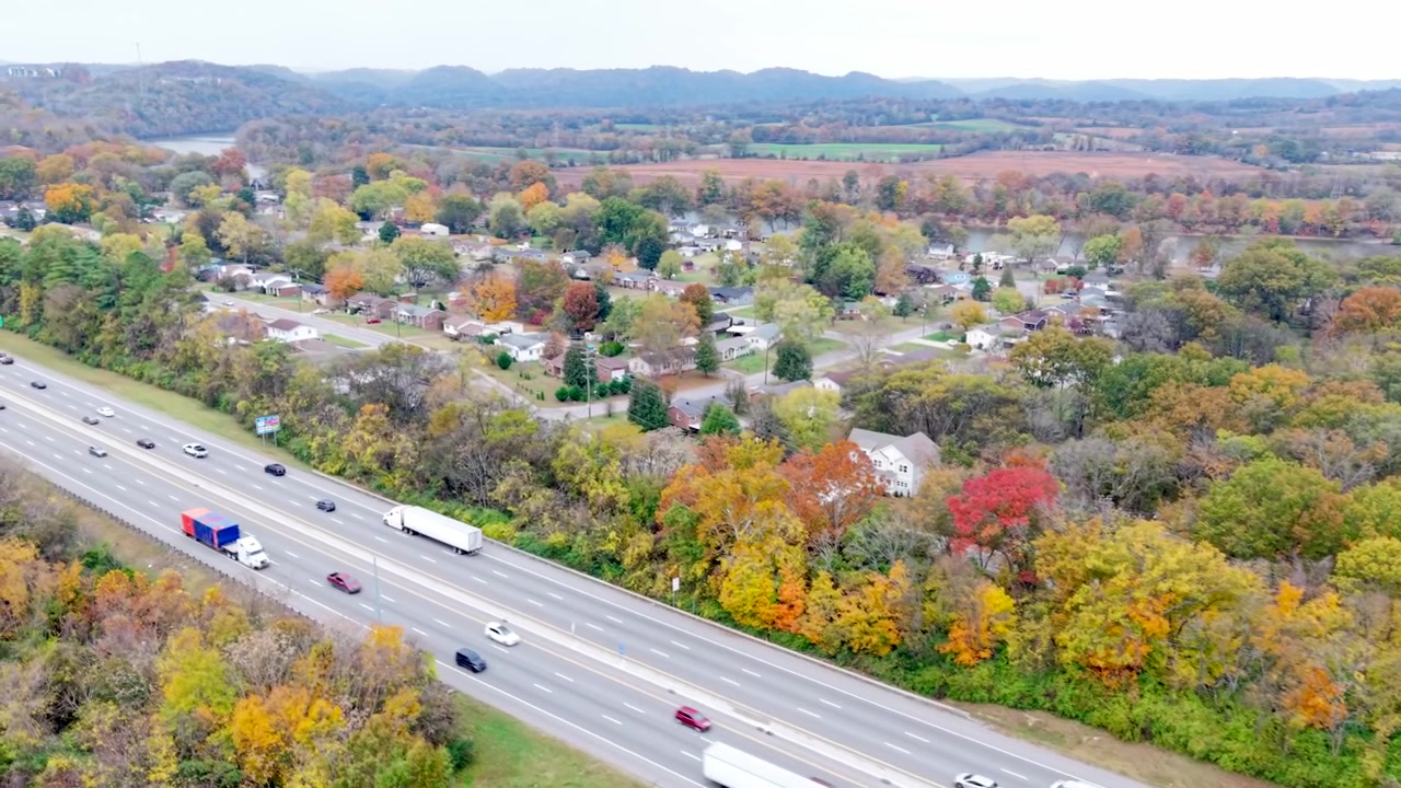 Un'autostrada trafficata delimitata da alberi autunnali colorati e case, che cattura paesaggi suburbani panoramici, traffico vivace e un vibrante fogliame autunnale in una morbida luce naturale - vista aerea scorrevole.
