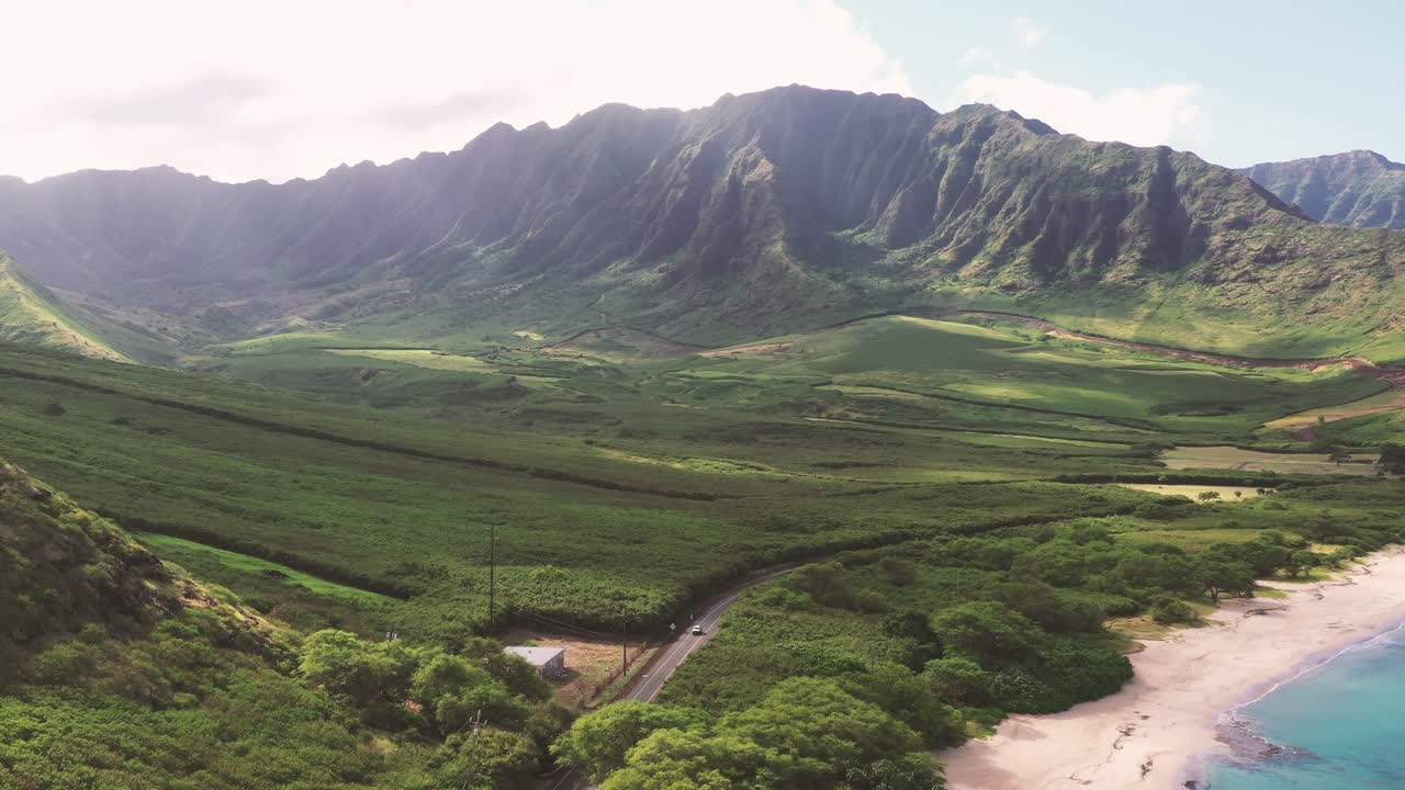 Vista aerea di una strada costiera che si snoda attraverso una vegetazione lussureggiante lungo una baia turchese. Le creste montuose illuminate dal sole di Ka'ena Point a Oahu creano uno sfondo mozzafiato a questo suggestivo paesaggio tropicale