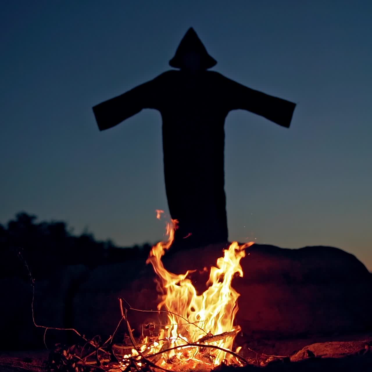 Black death behind the fire flame outdoors. Scary witch in black costume waving arms making fear on the background of burning fire. Halloween costume.