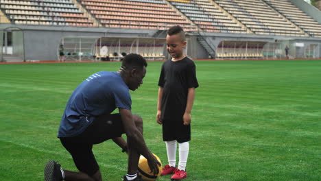 diverse trainer and child speaking on football field zoom in view of black male coach explaining game strategy to mixed race boy during football training on stadium