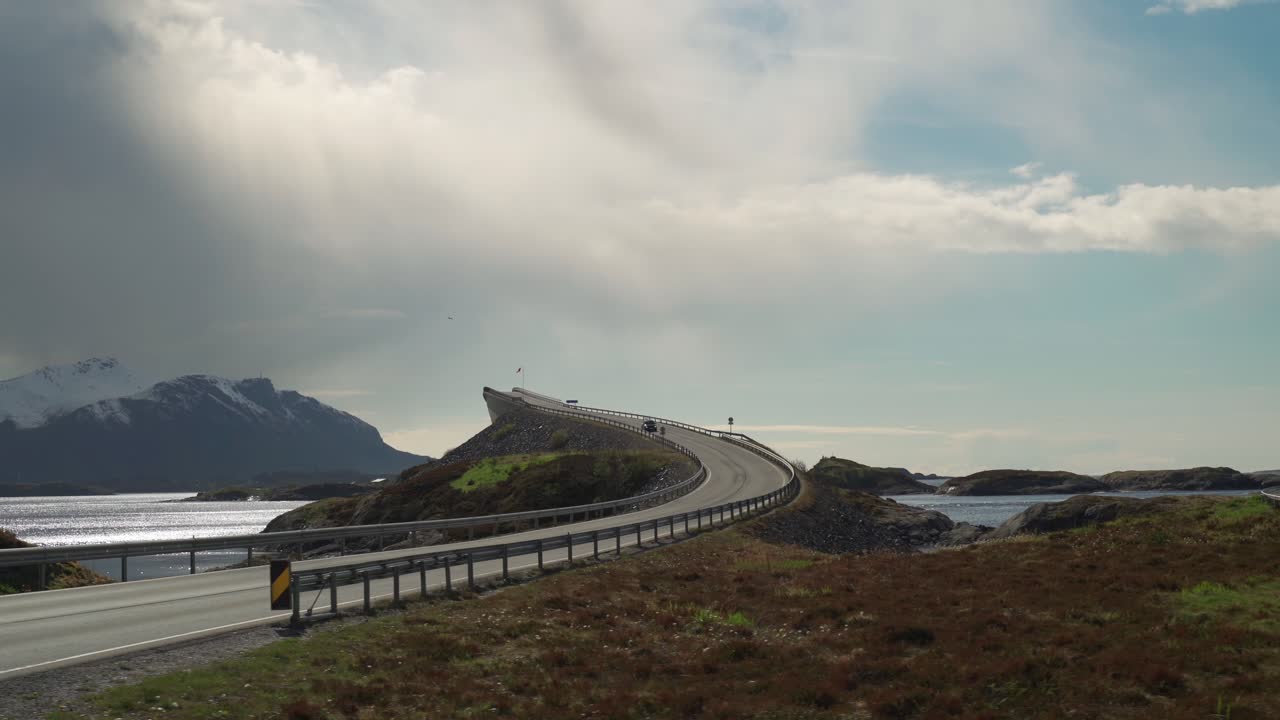 A famous Atlantic Road in Norway. One of its'  curved bridges rises above the sea. It is overcast, with heavy gray clouds hanging above the water. A dark silhouette of a mountain on the horizon