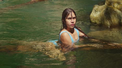 A-young-girl-with-Hispanic-heritage-relaxes-on-a-log,-wearing-a-bikini,-while-basking-in-the-serene-waters-of-a-crystal-clear-plunge-pool-at-a-picturesque-waterfall