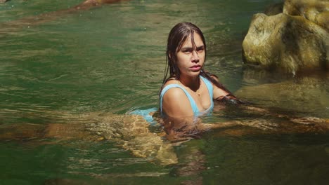 a young girl with hispanic heritage relaxes on a log, wearing a bikini, while basking in the serene waters of a crystal-clear plunge pool at a picturesque waterfall