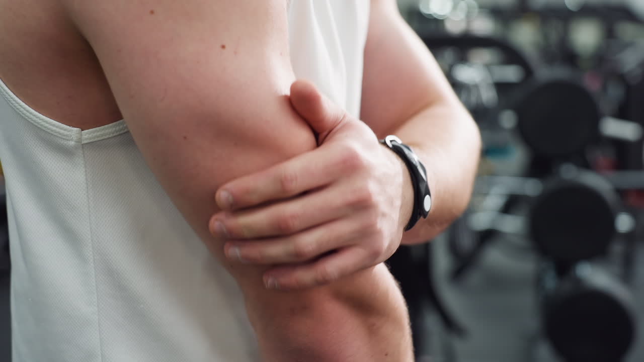 Body builder slaps hands against flexed arm muscles to loosen tension post workout, white singlet and black shorts visible, gym equipment blurred in background, focus on muscular definition
