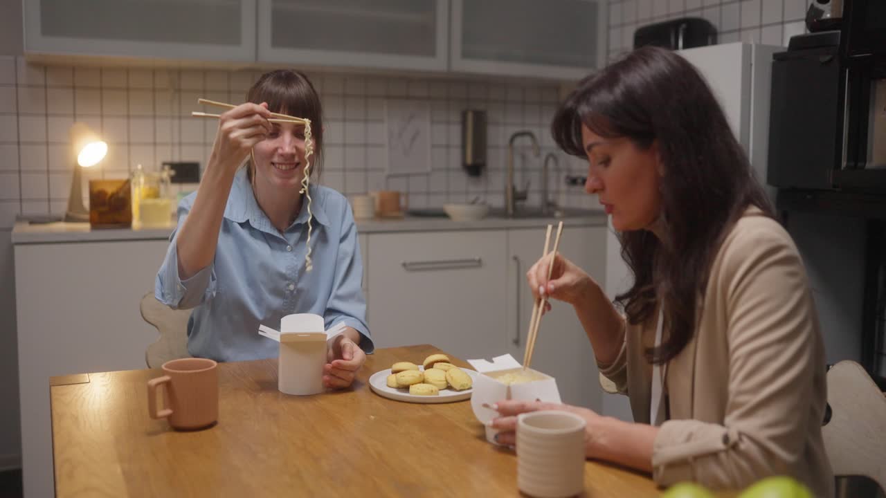 Two women eating takeout noodles and cookies in a kitchen