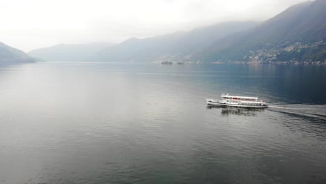 Aerial-view-of-a-boat-cruising-on-Lago-Maggiore-near-Ascona-in-Ticino,-Switzerland-with-view-of-the-Swiss-Alps-and-city-shore-in-the-background