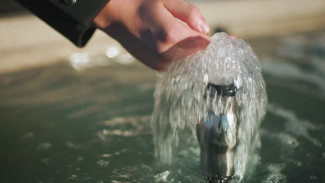 hand of man in black coat interacting with flowing spring water from small fountain, highlighting bubbling water surface, reflections, and dynamic movement in outdoor setting