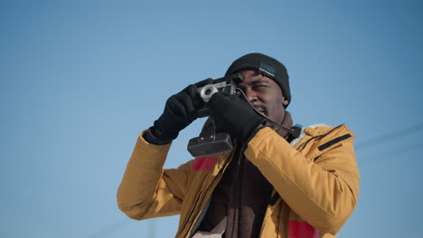 pressman carrying bag and camera as he fixes eyes on camera to take pictures in bright sun on busy urban sidewalk with pedestrians and traffic in winter scene adjusting vintage lens strap