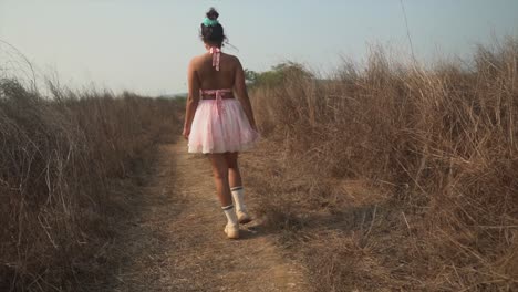 the back of a carefree asian female wearing a pink tutu dress walking along a remote path in a rural grass field on a hot summer’s day outdoors, india