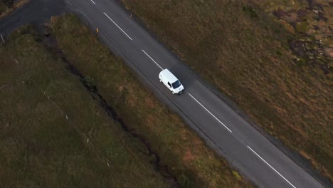 white car travels on small rural road with grass on sides, cloudy day, tracking aerial shot