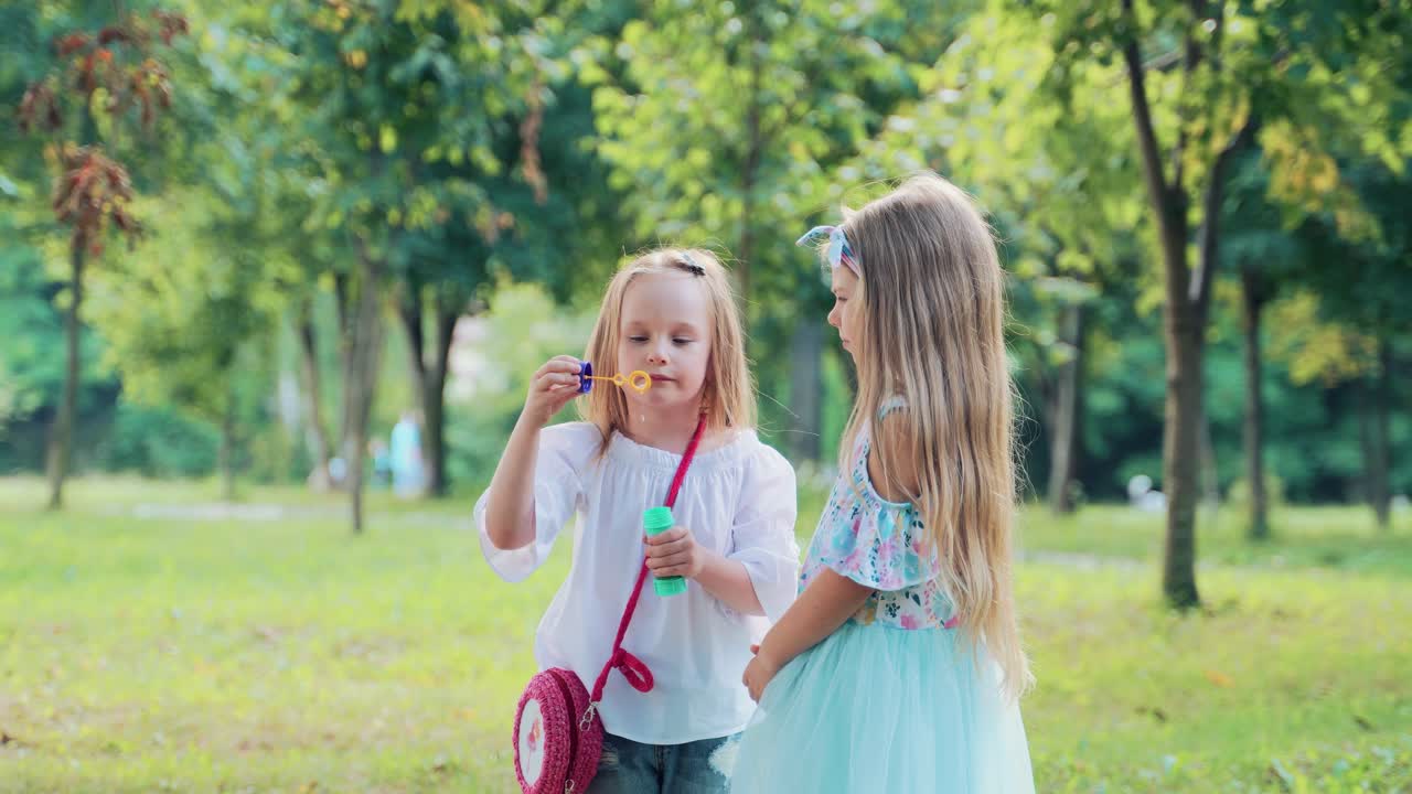 Little happy girls on a holiday in the park play, have fun and blow soap bubbles. Sunny summer day, green grass and trees